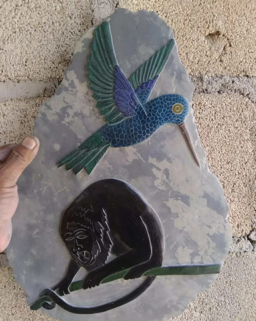 Stone Carvings of Humming Bird With Wings out Green Tips of Feathers, Body All Blue, Flying over Black Ape on Tree Limb